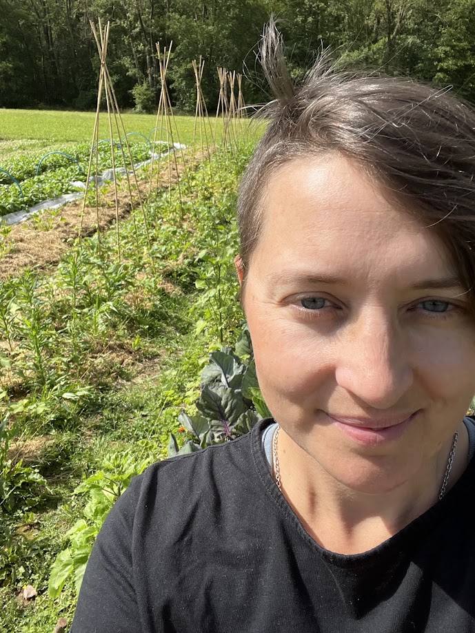 Selfie of Lindsey, head grower and founder of Llysiau Coed Cae. They are smiling in the sunshine, the background shows rows of plants growing.